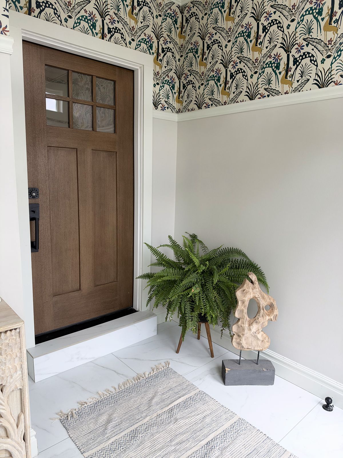 The original wood front door framed by botanical wallpaper, with a fern, a found-wood sculpture and a woven rug on marble-look tile