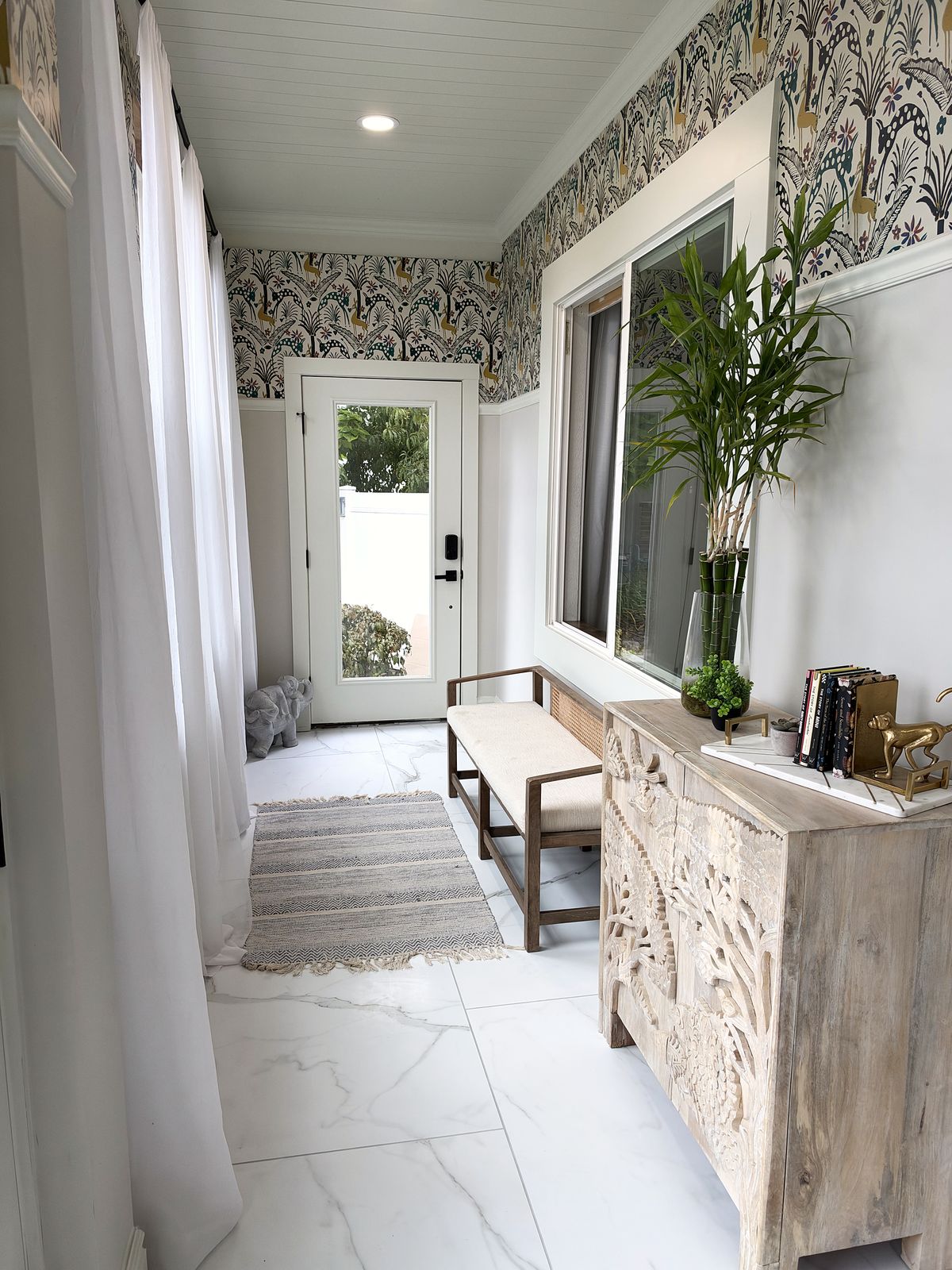 Enclosed porch mudroom with botanical wallpaper above white wainscoting, marble-look tile, a carved wood console, a bench and sheer curtains