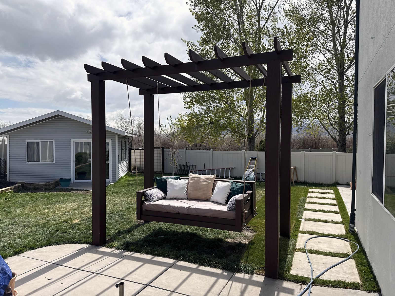 Dark-stained pergola with a hanging daybed swing piled with cushions, beside a path of concrete stepping pads