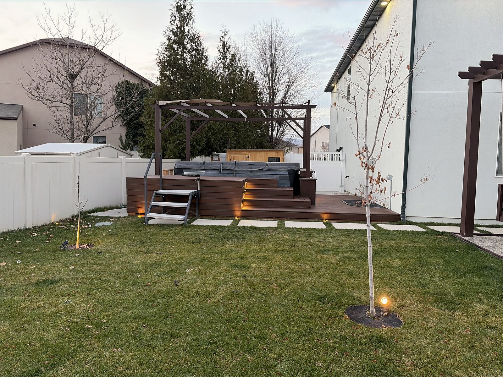 Backyard at dusk with an elevated stained-wood deck, a covered hot tub under a pergola, stepping pads across the lawn and low landscape lighting